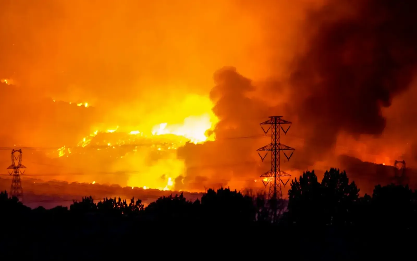 Wildfire burning on a hillside at night behind power-line towers